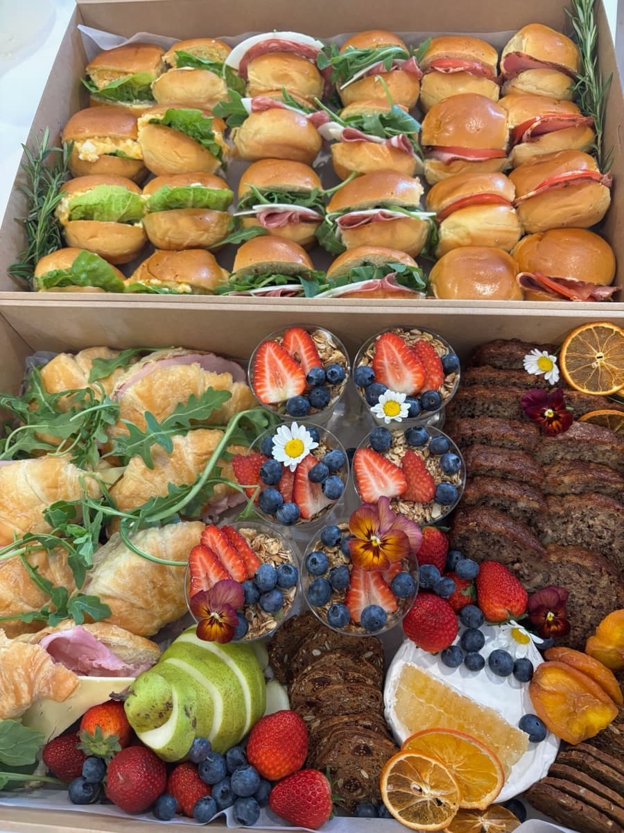 Brunch catering spread with egg and bacon sliders, ham and cheese rolls, golden croissants, granola parfait cups with berries, banana bread, brie, and seasonal fruit