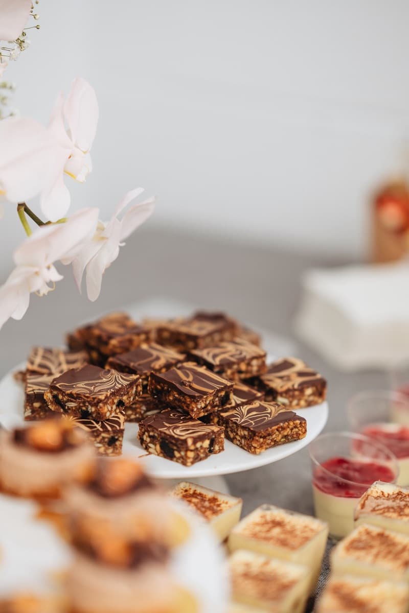 Chocolate and peanut butter brownie slices with marbled ganache topping on a white cake stand, with tiramisu cups and berry cups visible alongside, for Adelaide corporate dessert catering