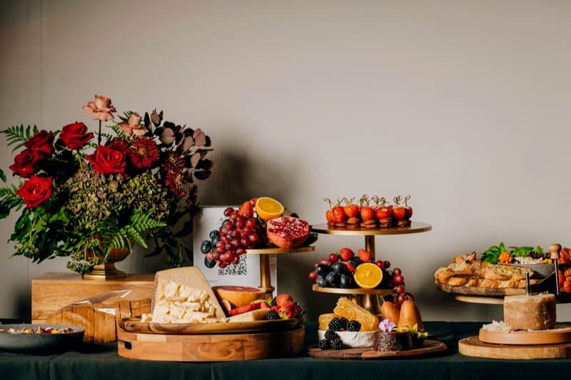 Festive grazing table with red roses, gold tiered stands, pomegranate, seasonal fruit, and charcuterie