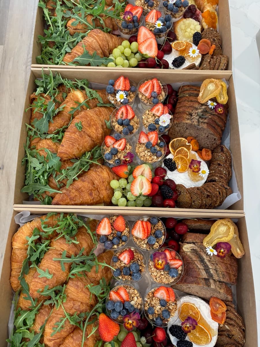 Three Event Gourmet brunch boxes packed with golden croissants, granola parfait cups, banana bread, brie, fresh berries, grapes, and edible flowers