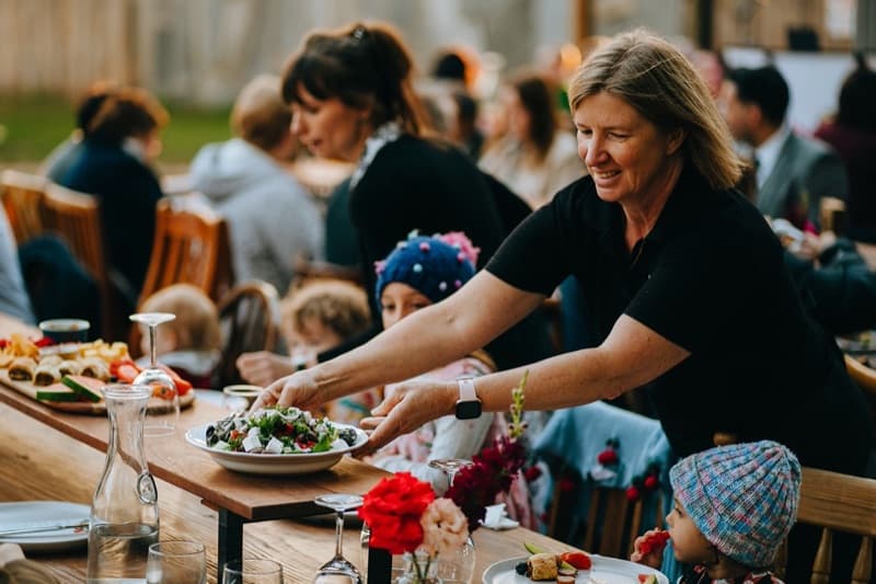Rose serving plated dishes to guests at a long timber dining table at an outdoor Adelaide wedding reception